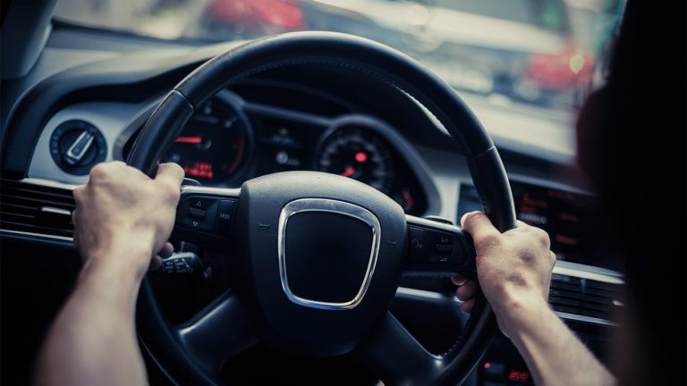 Driver gripping the steering wheel, viewed from inside the cabin with dashboard instruments illuminated