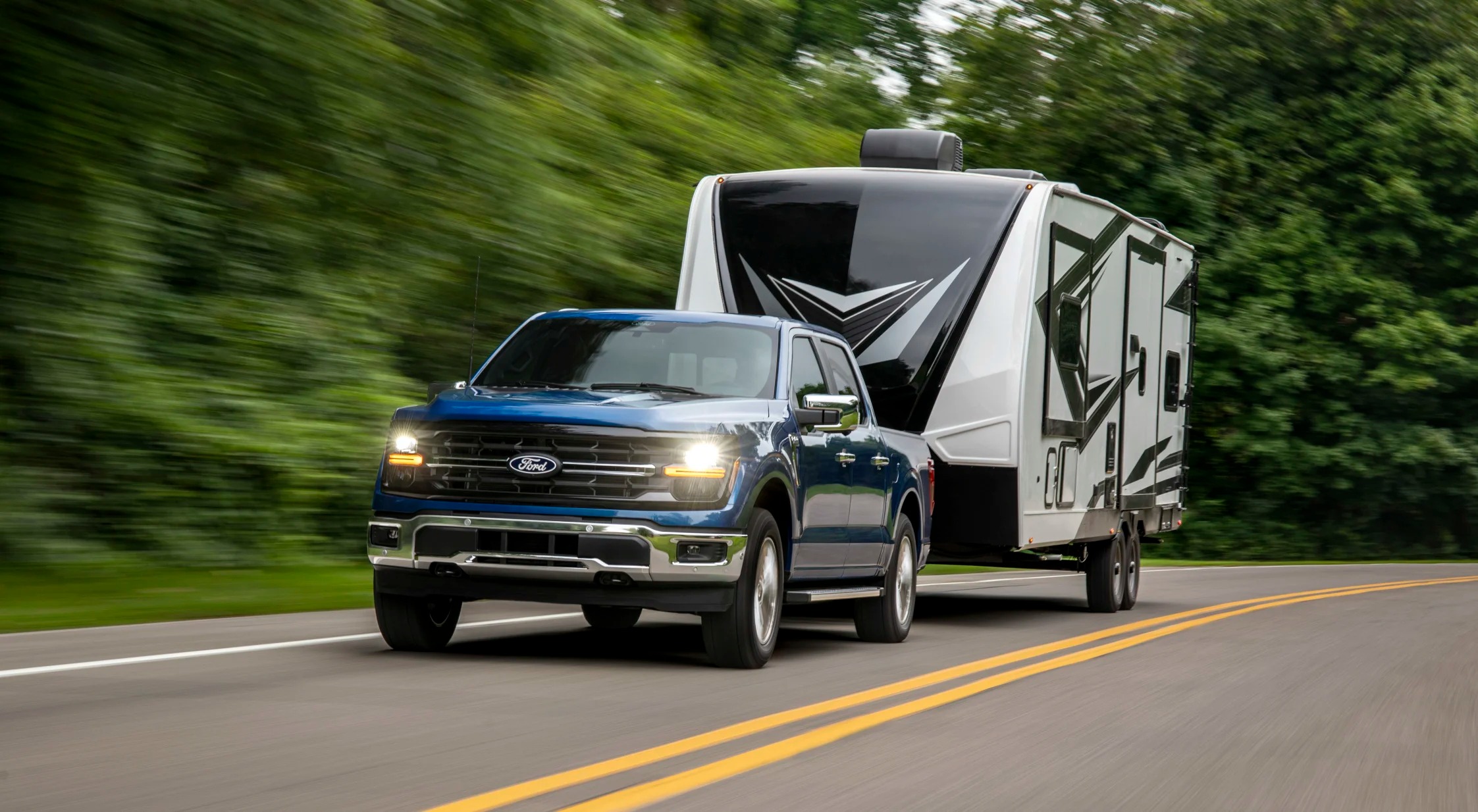Ford F-150 tows a large fifth-wheel camper along a tree-lined highway