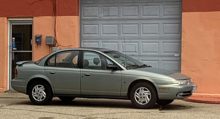 Green Saturn S-Series sedan parked outside an orange building, showcasing its classic 1990s design