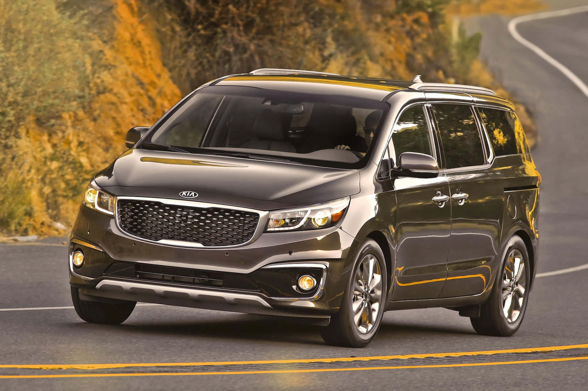Kia Sedona minivan driving along a winding road, showcasing its spacious family-friendly design and illuminated headlights