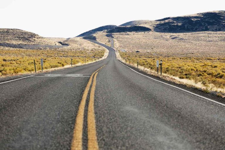 Empty highway stretches through dry desert landscape with distant hills