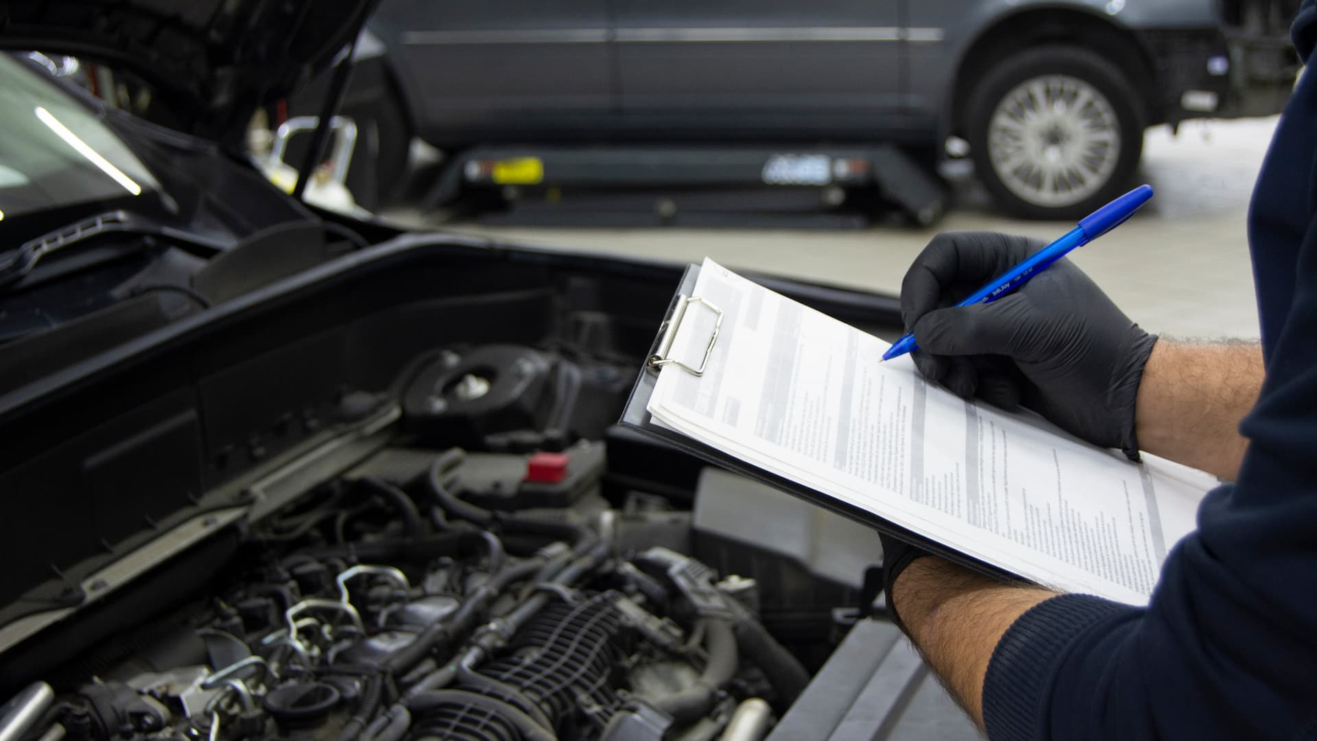 Mechanic documents vehicle inspection findings while examining an open engine bay in an automotive workshop