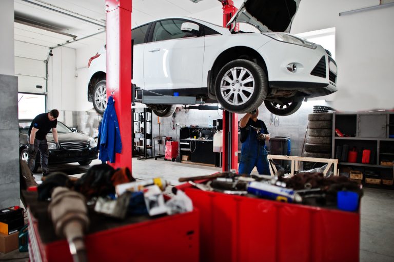 Mechanics inspect a raised car inside an automotive repair workshop, with tools and spare parts nearby