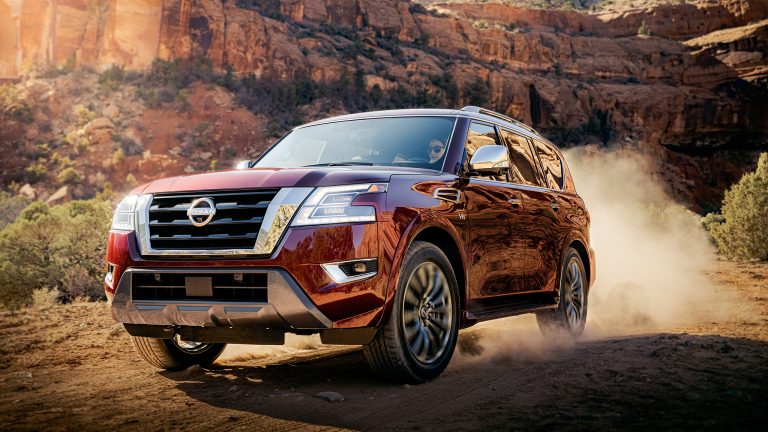 Nissan Armada driving across rugged desert terrain, kicking up dust beneath towering red rock cliffs