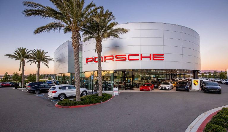 Porsche dealership featuring a modern curved showroom, prominent red signage, palm trees, and luxury sports cars
