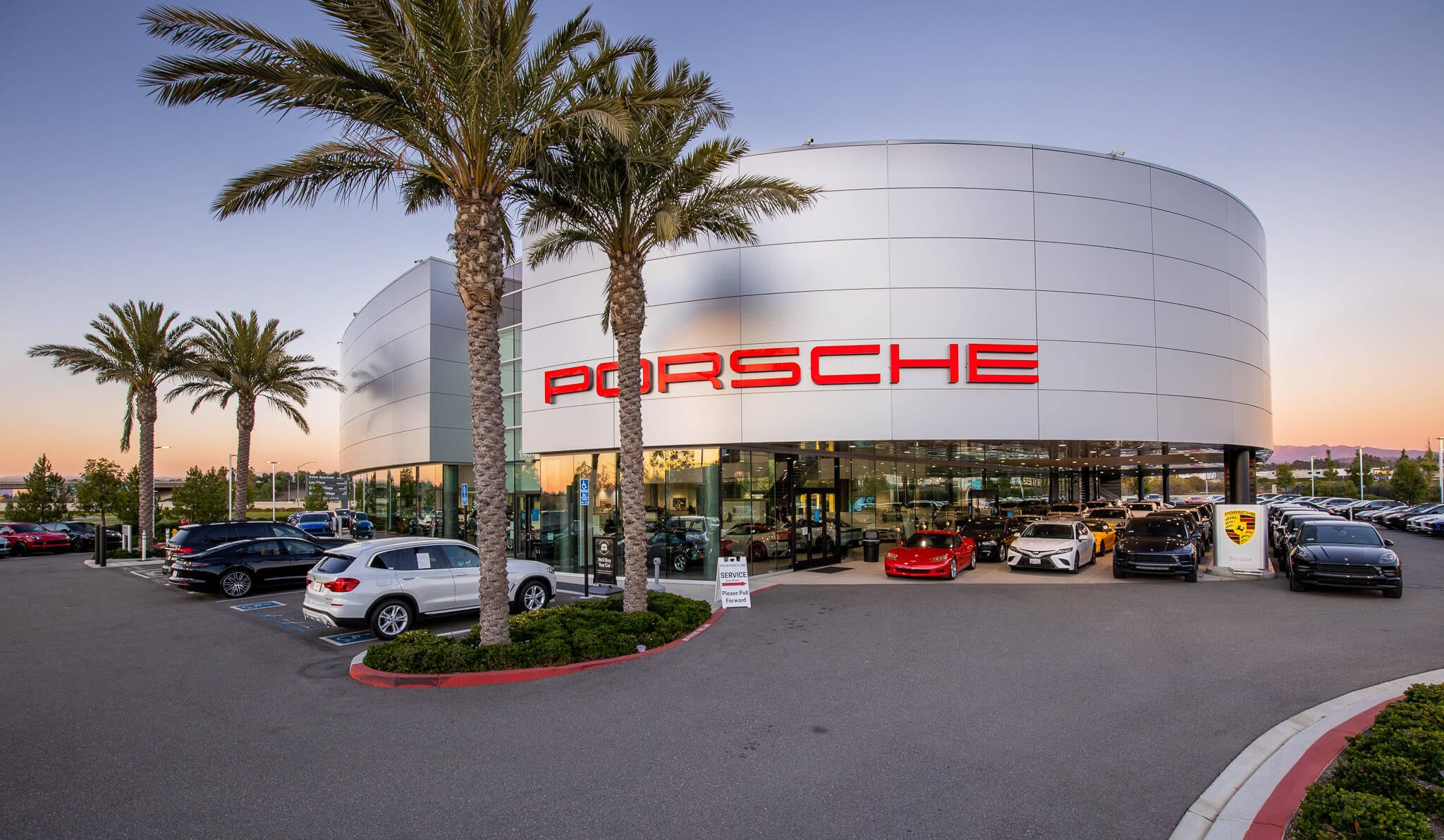 Porsche dealership featuring a modern curved showroom, prominent red signage, palm trees, and luxury sports cars