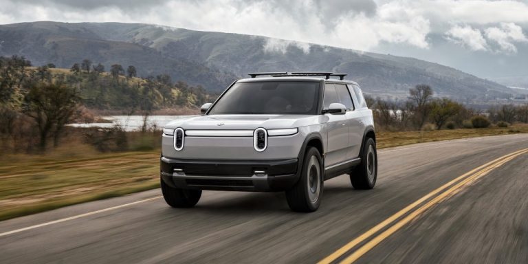 Rivian R2 electric SUV driving along a scenic mountain road beneath an overcast sky
