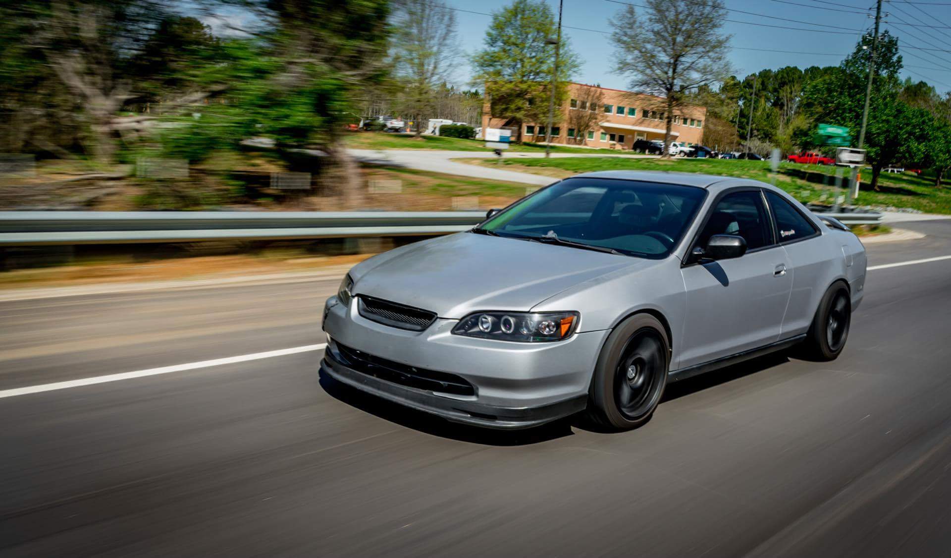 Silver Acura RSX driving along a road, showcasing its sporty coupe styling and black alloy wheels