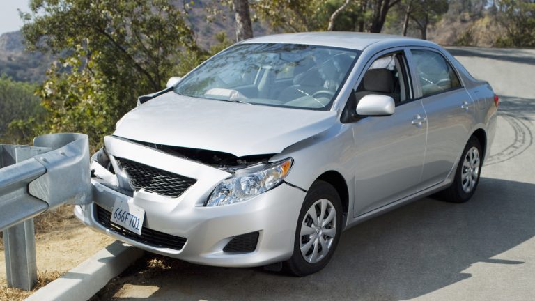 Silver sedan with significant front-end collision damage resting against a roadside metal guardrail