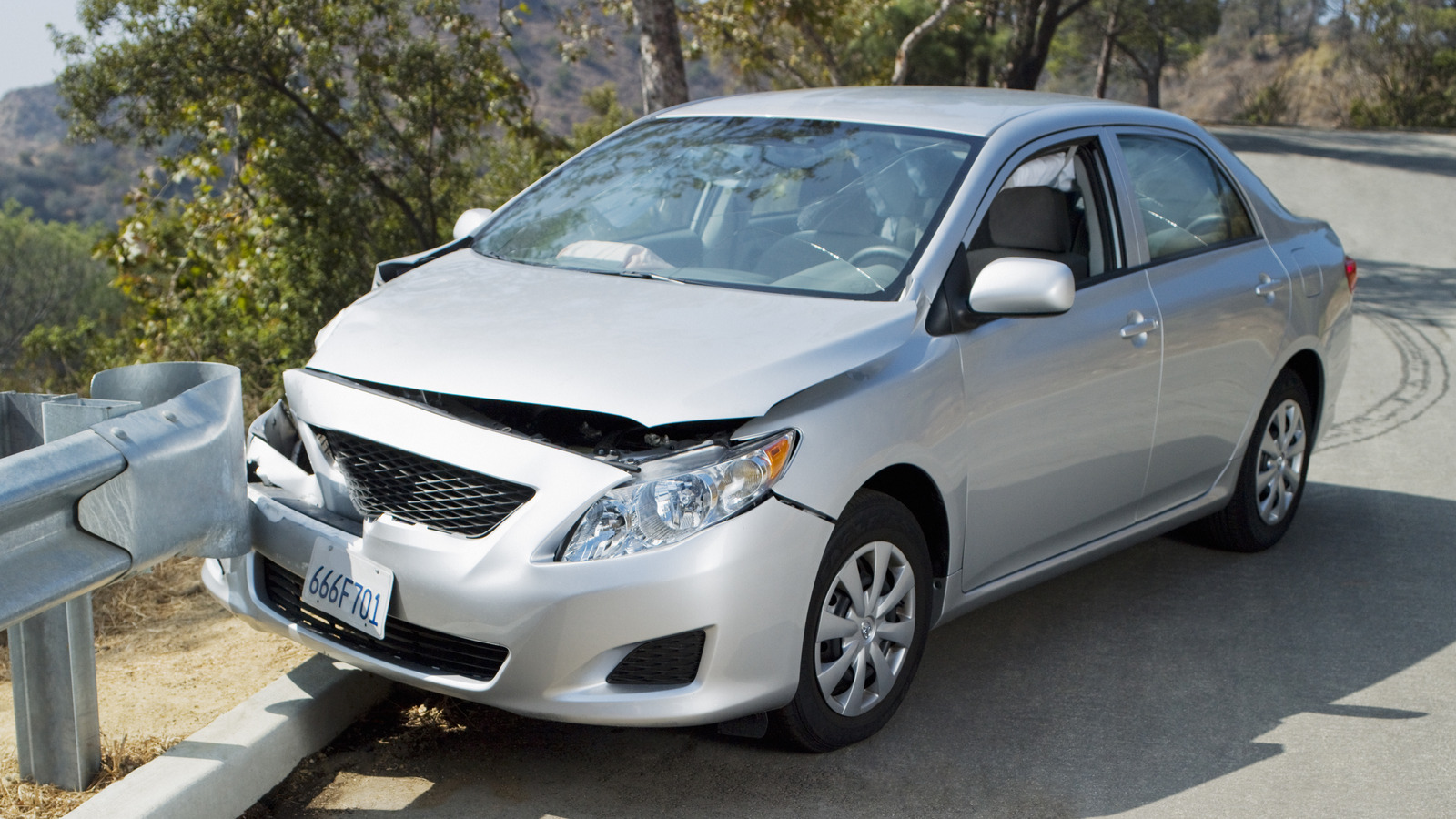 Silver sedan with significant front-end collision damage resting against a roadside metal guardrail