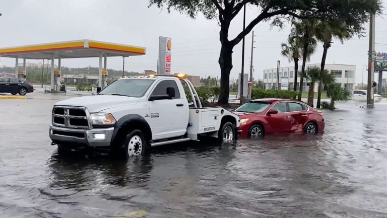 Tow truck assists a stranded red sedan stranded in deep floodwater near a gas station