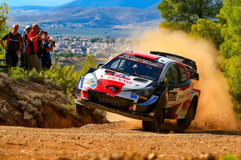 Toyota Yaris rally car launches over rugged terrain, scattering dust as spectators watch nearby