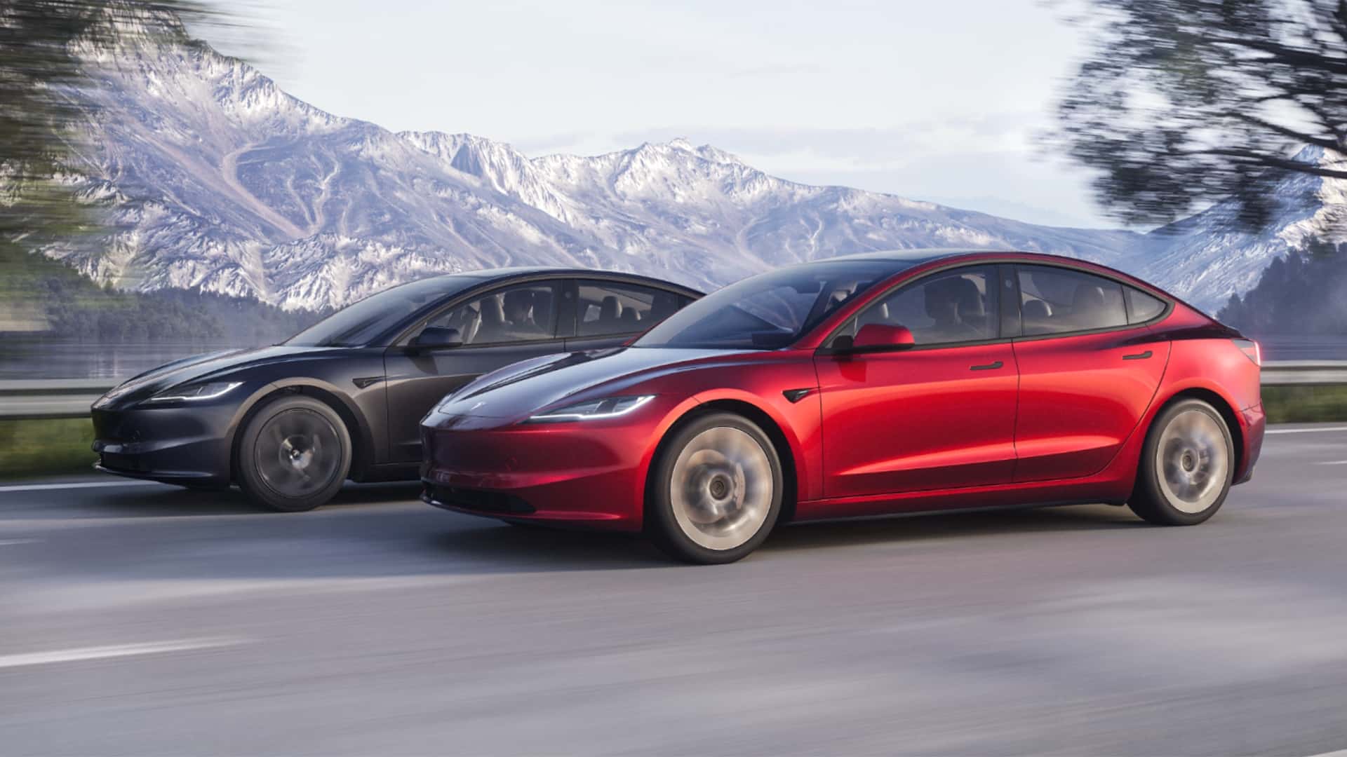 Two Tesla Model 3 sedans drive along a scenic highway beneath snow-capped mountains