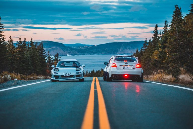 Two sports cars meet on a scenic mountain road beside a lake at dusk