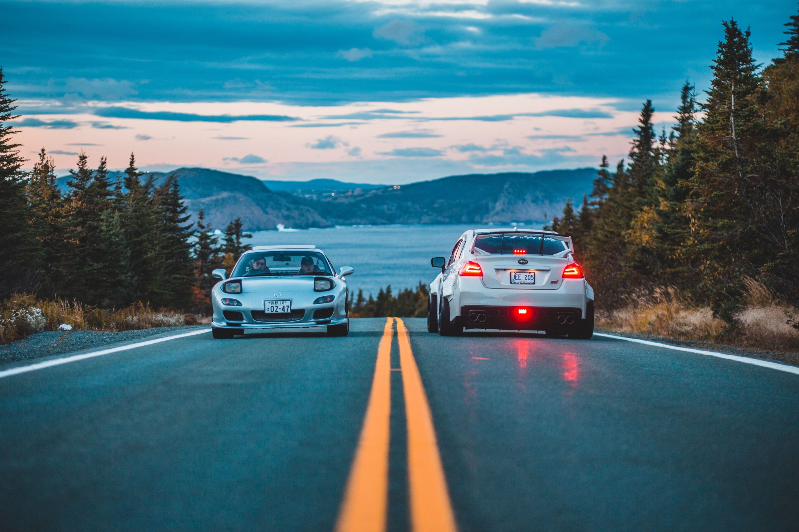 Two sports cars meet on a scenic mountain road beside a lake at dusk