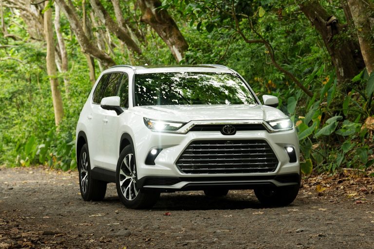 White Toyota Grand Highlander photographed on a wooded road, showcasing its bold front grille and spacious SUV design