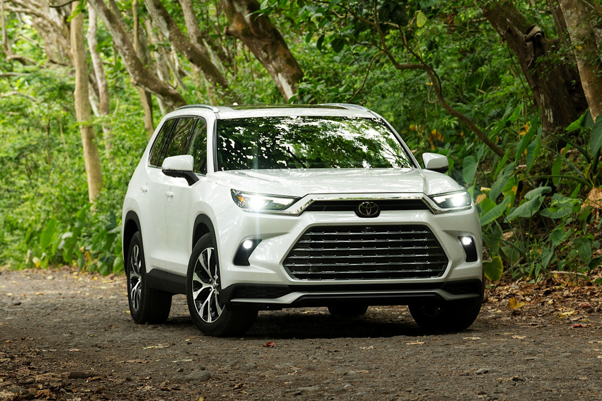 White Toyota Grand Highlander photographed on a wooded road, showcasing its bold front grille and spacious SUV design