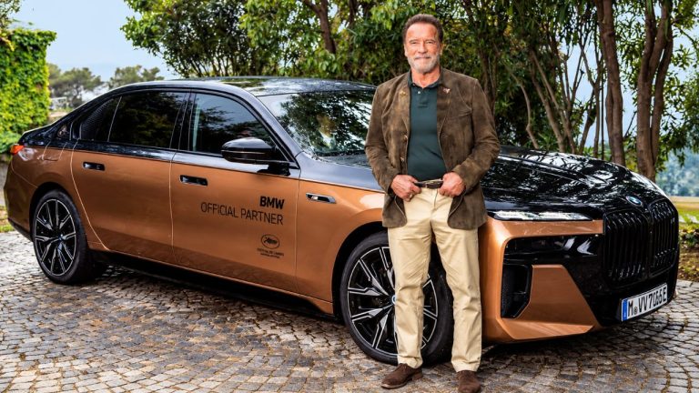 Arnold Schwarzenegger stands beside a luxury BMW sedan during an outdoor promotional event
