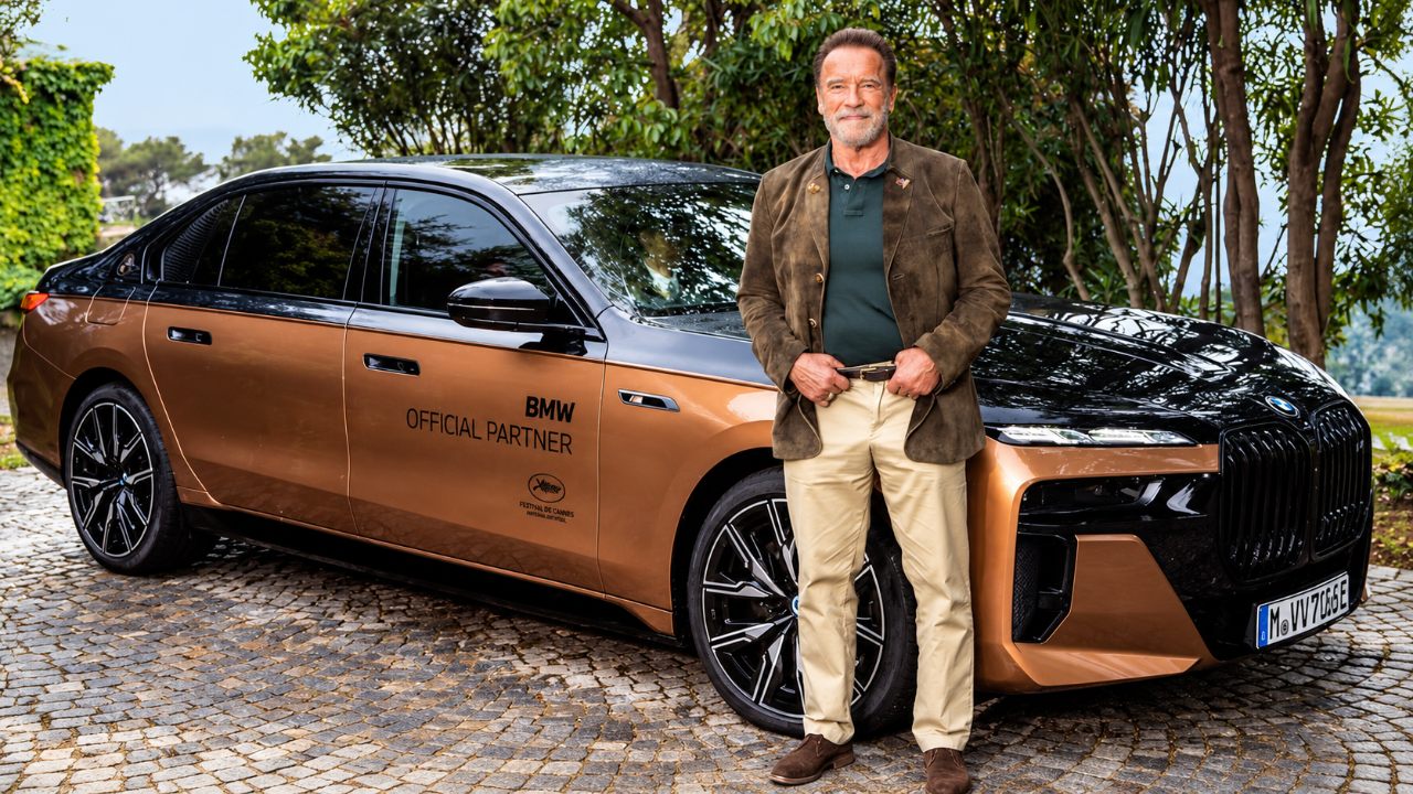 Arnold Schwarzenegger stands beside a luxury BMW sedan during an outdoor promotional event