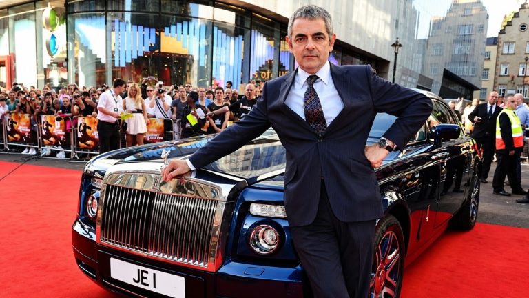 Rowan Atkinson poses beside a luxury Rolls-Royce on a crowded red carpet